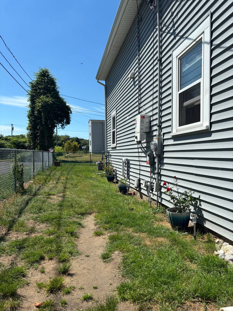Side view of a gray and white striped mobile home with foundation piers, surrounded by overgrown grass and weeds on a sunny day