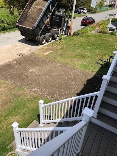 Dump truck unloading gravel onto residential driveway, view from deck with white railings